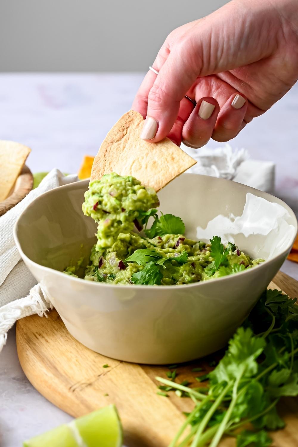 A bowl of homemade chipotle guacamole garnished with parsley served on a wooden cutting board. A hand is dipping a tortilla chip in it.