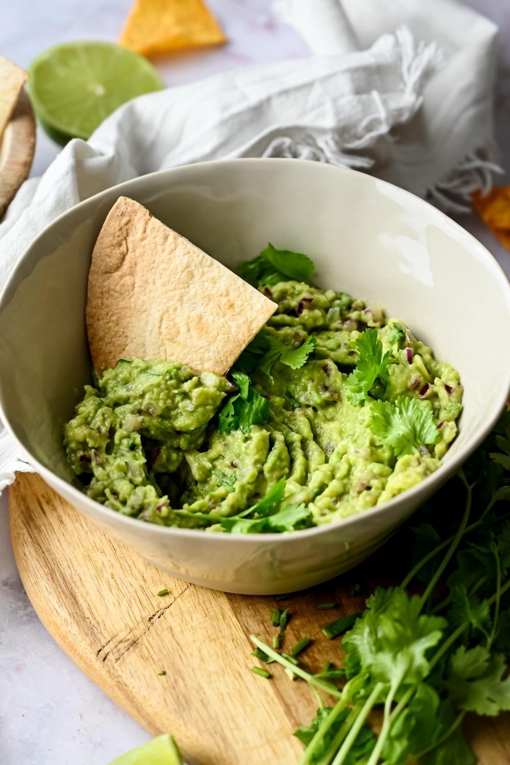 A bowl of homemade chipotle guacamole garnished with parsley served on a wooden cutting board. A tortilla chip is dipped in the bowl.
