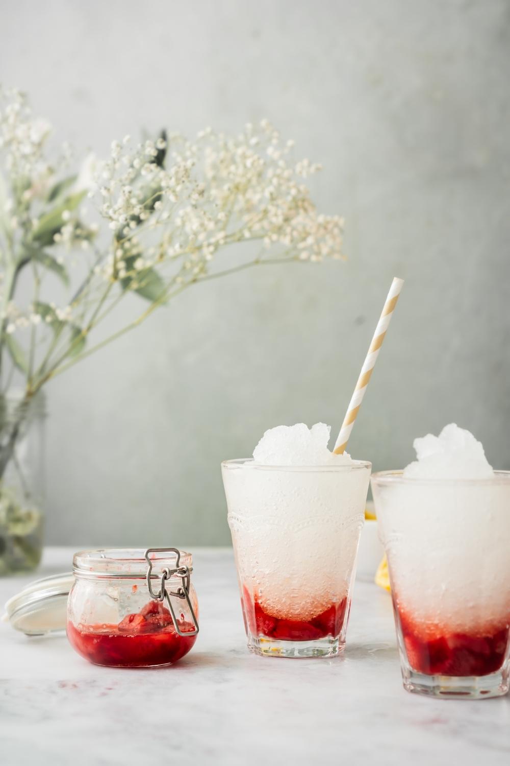 Two strawberry slushies in clear glasses, one with a straw sticking out. There is a small container of strawberries next to the two glasses.