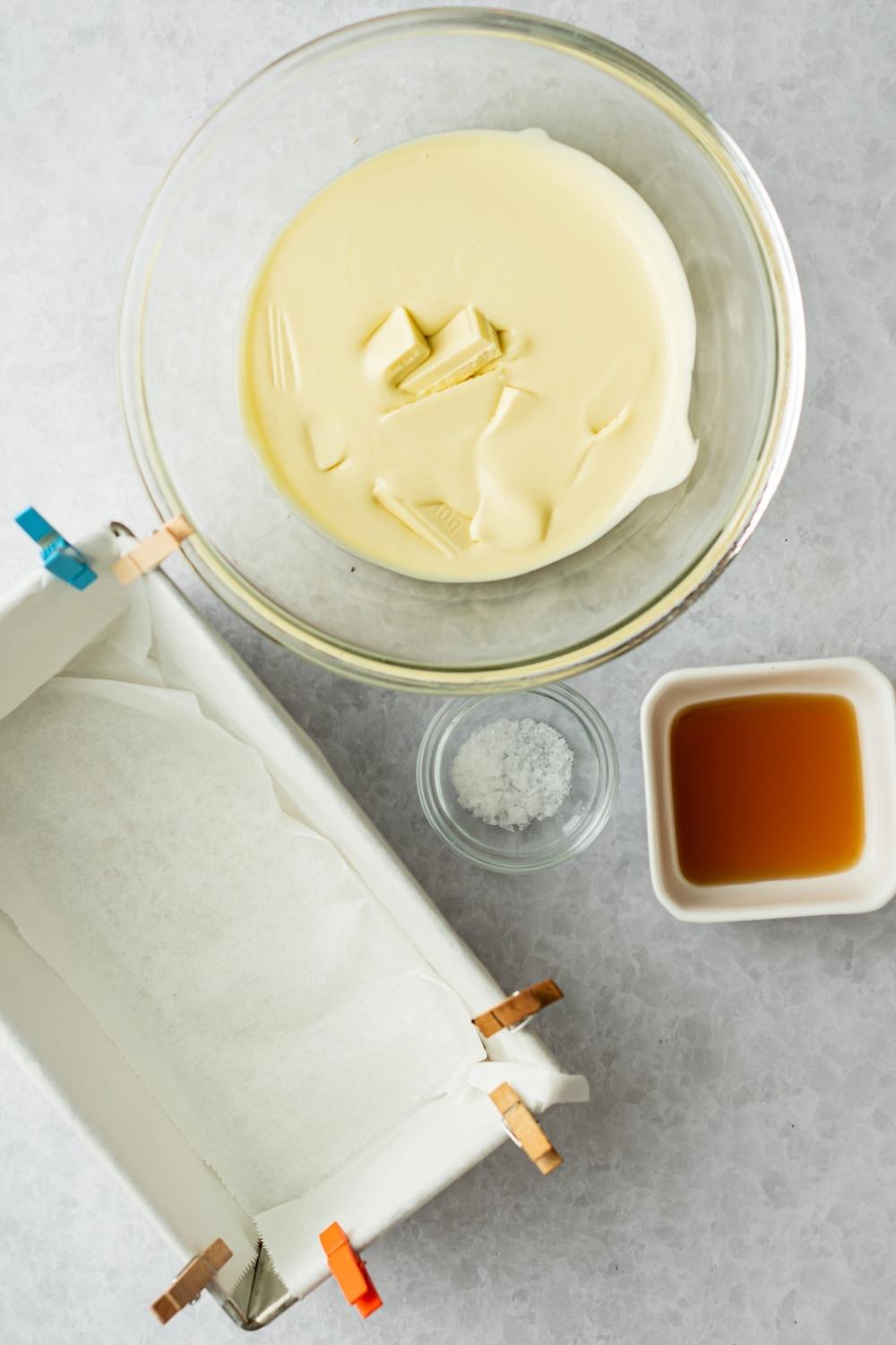 An overhead view of a loaf pan lined with parchment paper, a small bowl of maple syrup, a small bowl of salt, and a medium bowl with melted white chocolate.