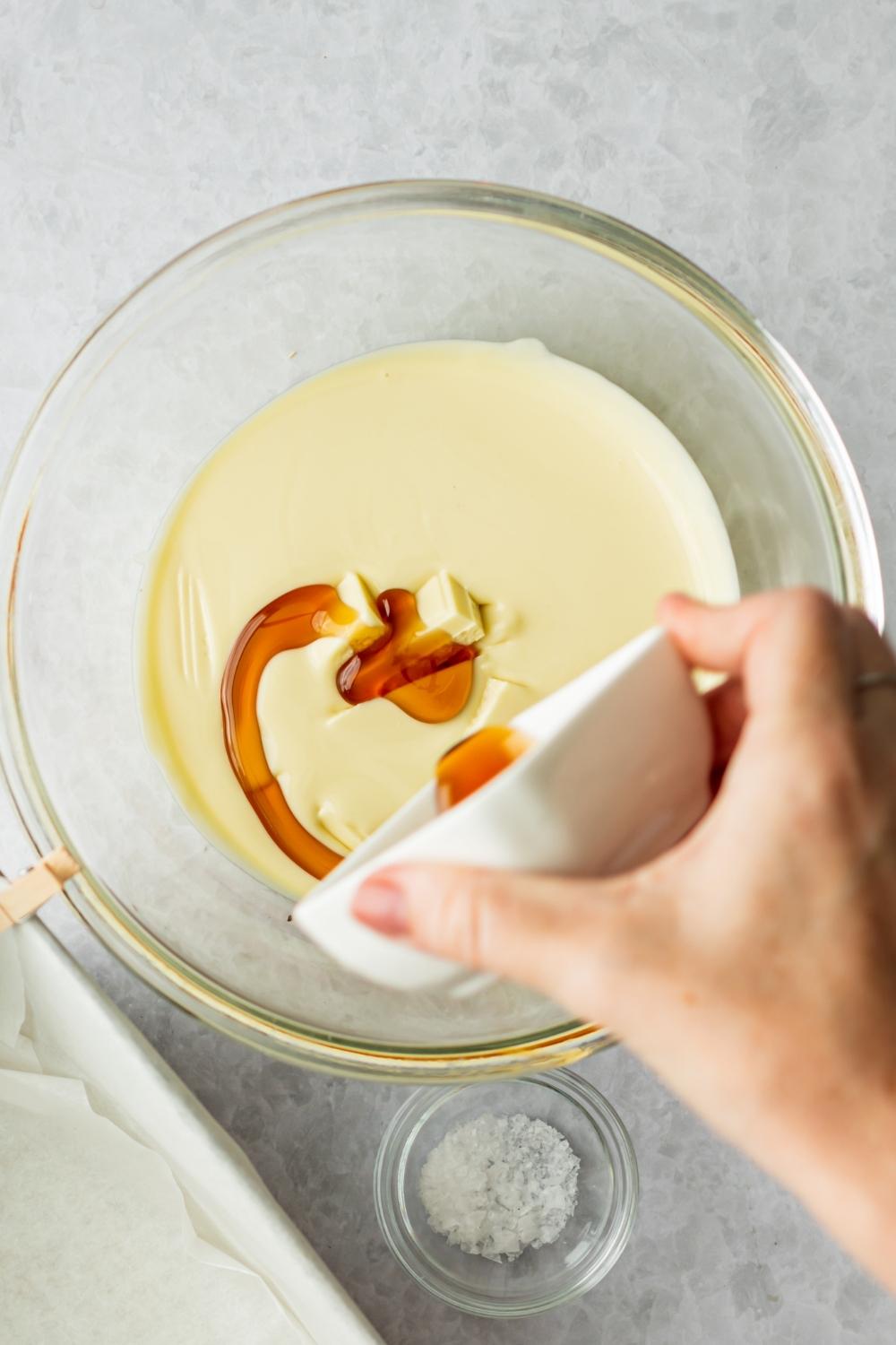 A medium bowl with melted white chocolate and a hand holding a small bowl pouring pure maple syrup into it.