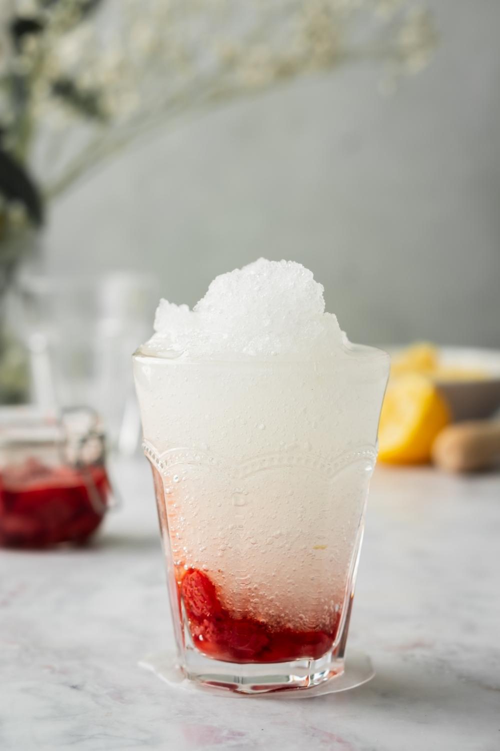 A finished strawberry slushie in a glass, with the ingredients to make the slushie behind the glass.