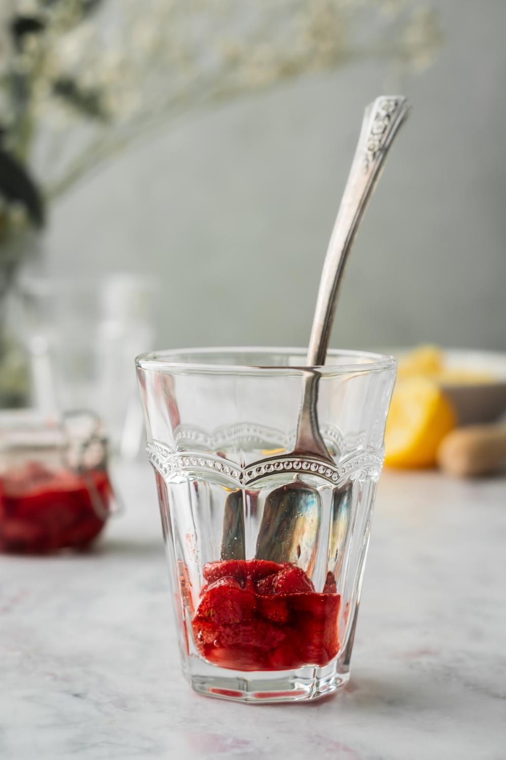 A cup with muddled strawberries and a spoon sticking out of the glass and ingredients to make a slushie behind the glass.