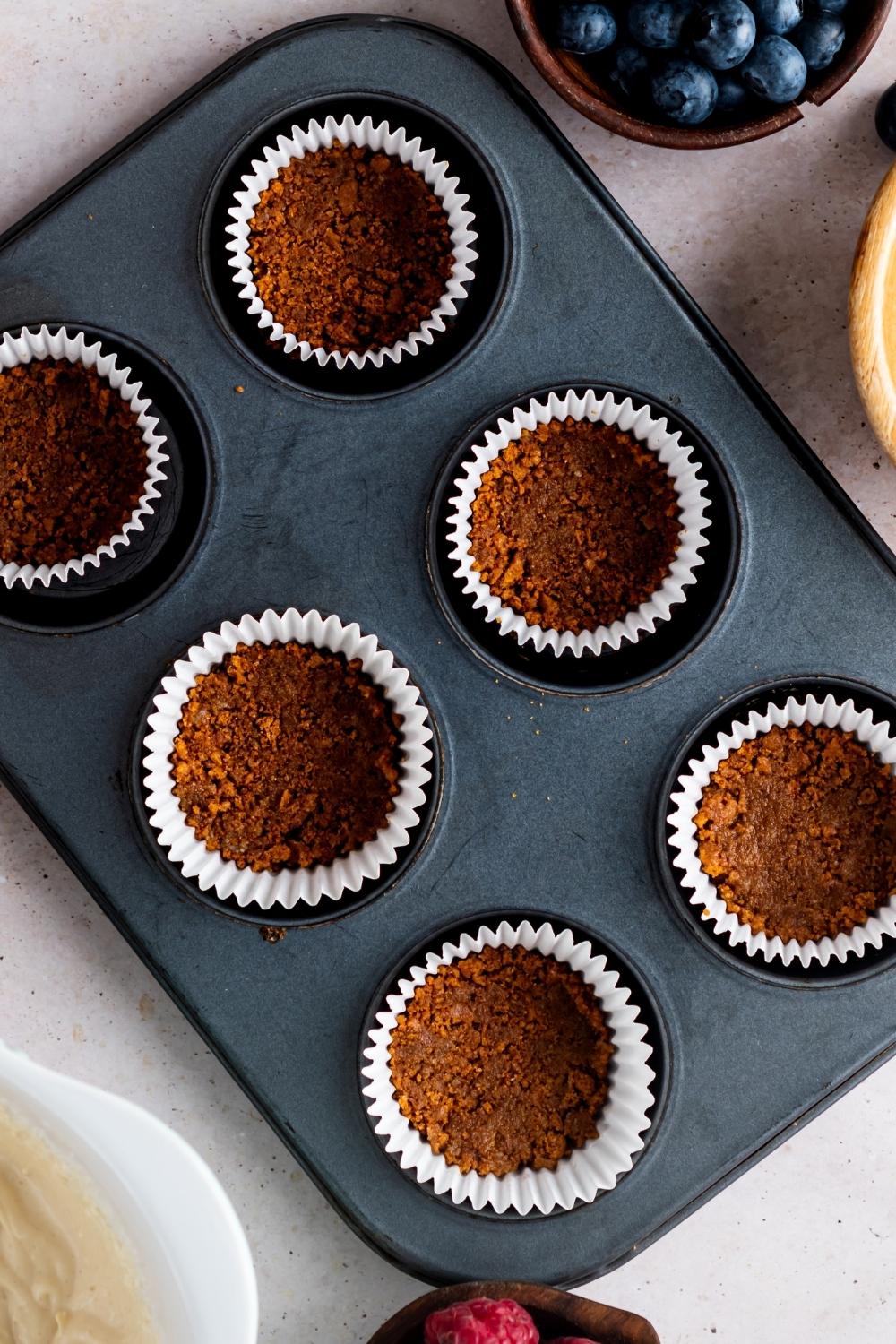 A muffin pan with graham cracker crust pressed in each muffin liner.