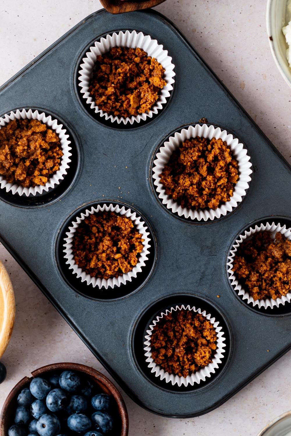 A muffin pan with graham cracker crust scooped in each muffin liner.