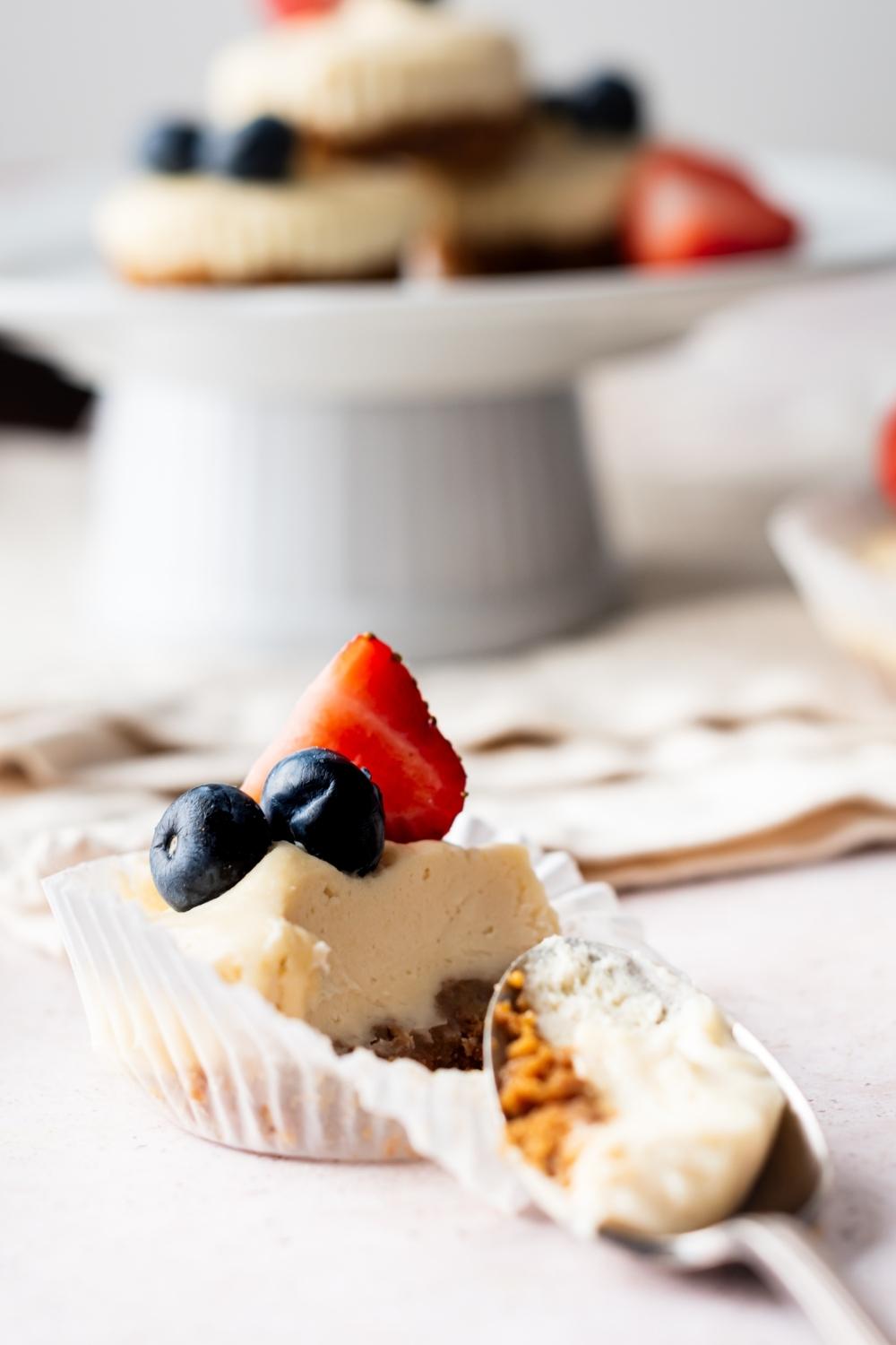 A mini cheesecake bite with a spoon sitting by it with a scoop of cheesecake in it. A cake stand with homemade mini cheesecake bites sits behind it.