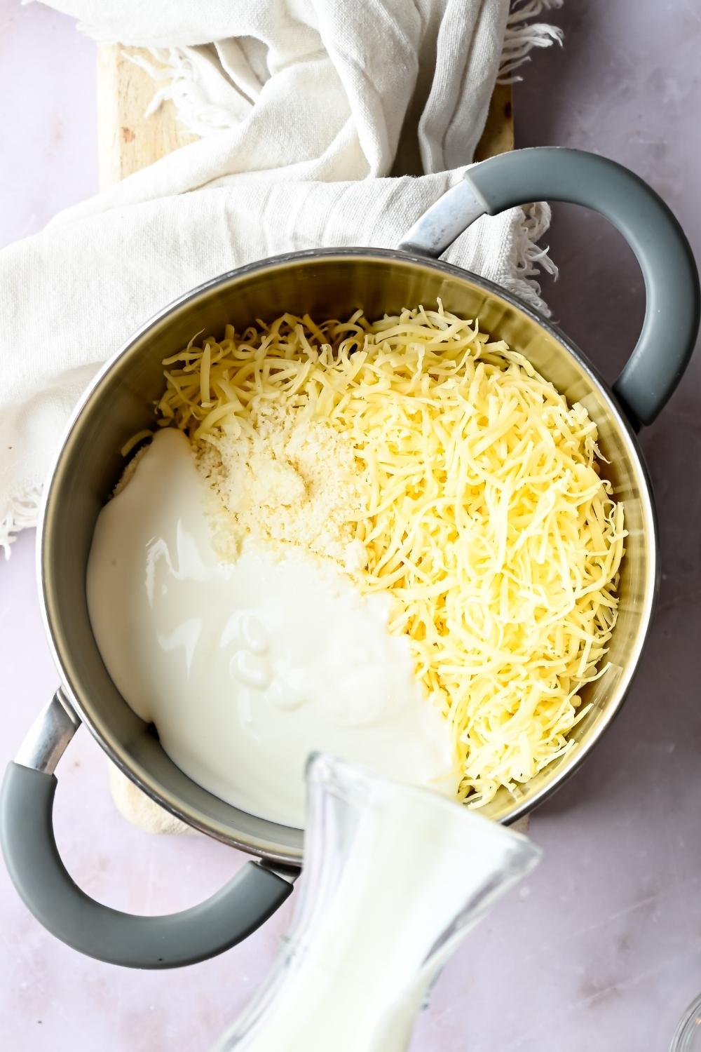 Overhead view of a medium pot with a blend of cheeses. A jar of half & half is being poured into it.