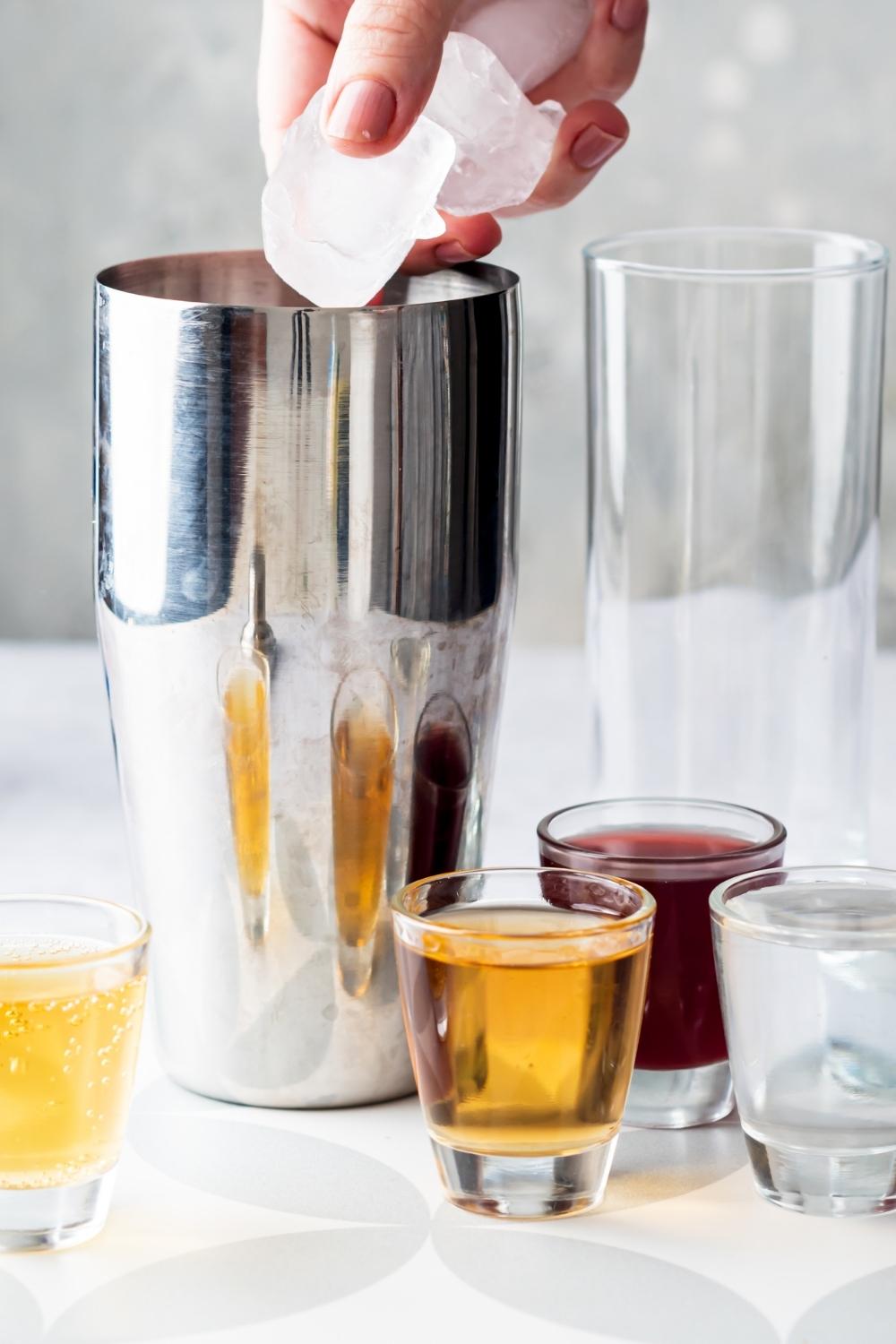 A cocktail shaker on a countertop next to an empty glass. Shot glasses it on the countertop around the shaker containing alcohol to make a Vegas bomb shot. A hand is dumping ice cubes into the shaker.