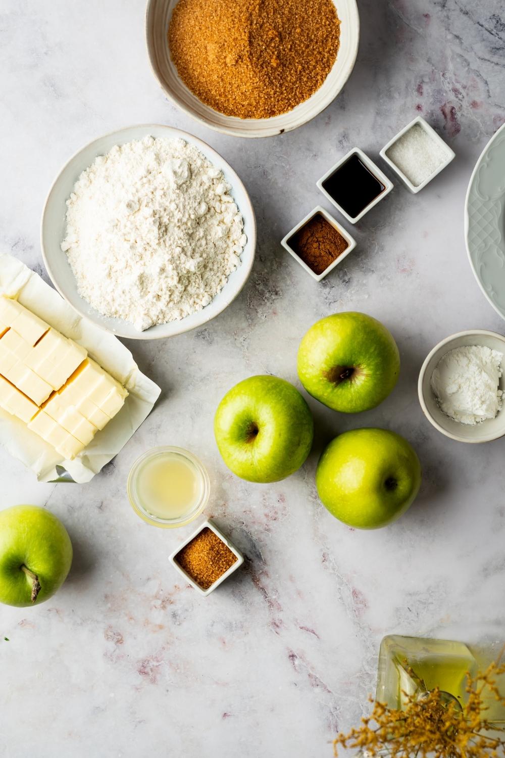 Four apple, a bowl of flour, cubed butter, and a bowl of brown sugar all on a grey counter.