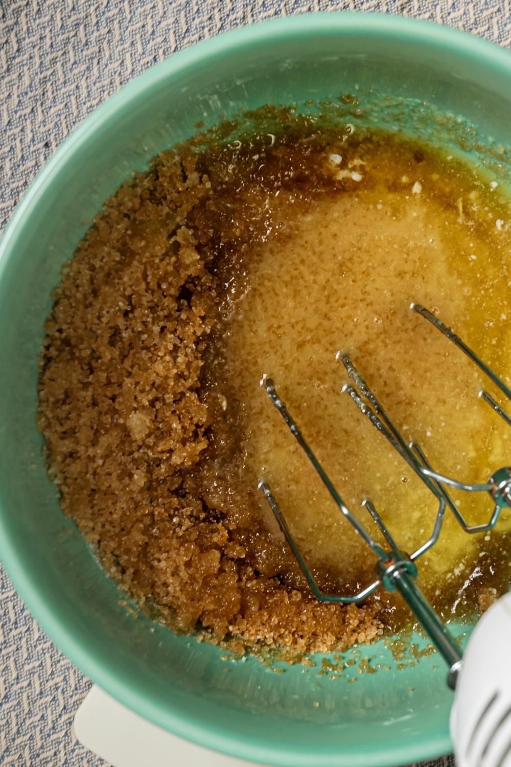 An overhead view of melted butter and brown sugar in a bowl. An electric mixer is in it.