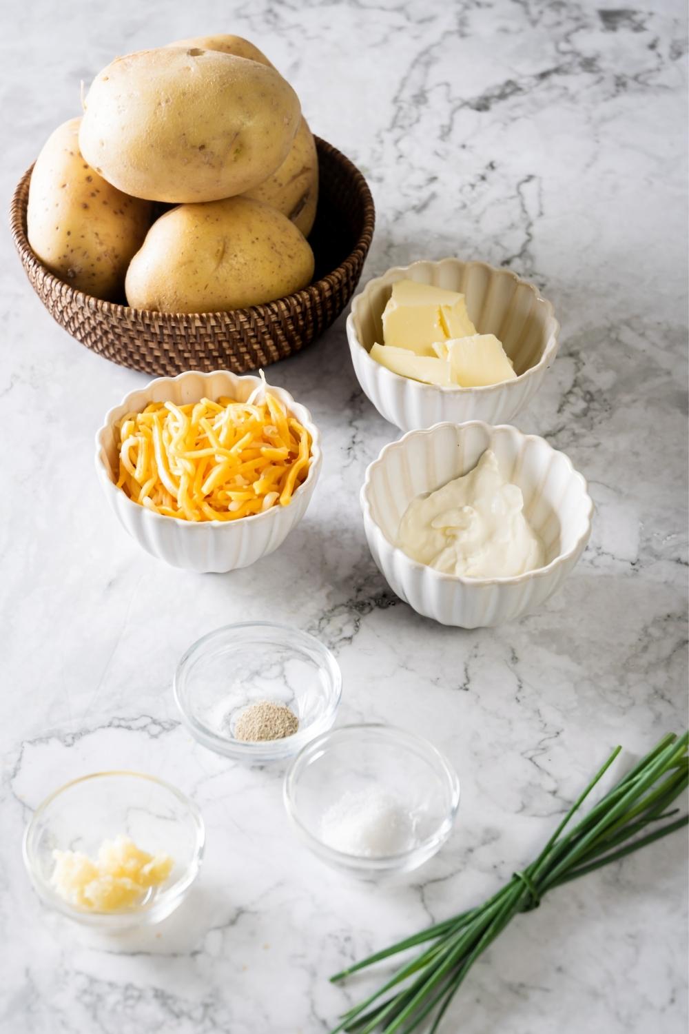 A bowl of potatoes, a bowl of cheddar cheese, a bowl of butter, a bowl of sour cream, a small bowl of salt, and a small bowl of pepper all on a white counter.
