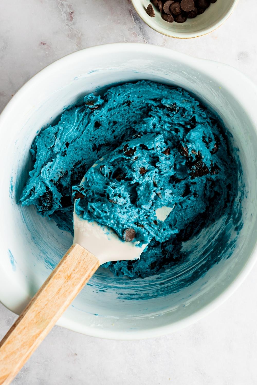 A mixing bowl of cookie monster cookie dough. Chocolate chips and Crushed oreos have been added to the dough. A rubber spatula sits in the bowl.