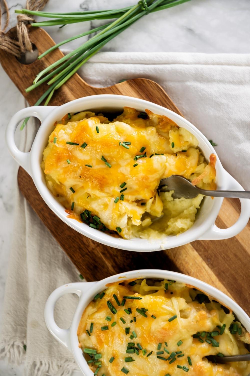 Cheesy mashed potatoes in a white casserole dish on a wooden cutting board. There is another casserole dish with the mashed potatoes in it in front of it.