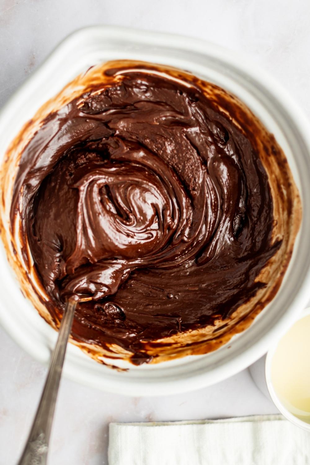 A mixing bowl containing melted chocolate and the ingredients needed for fireball fudge a spoon is mixing it.