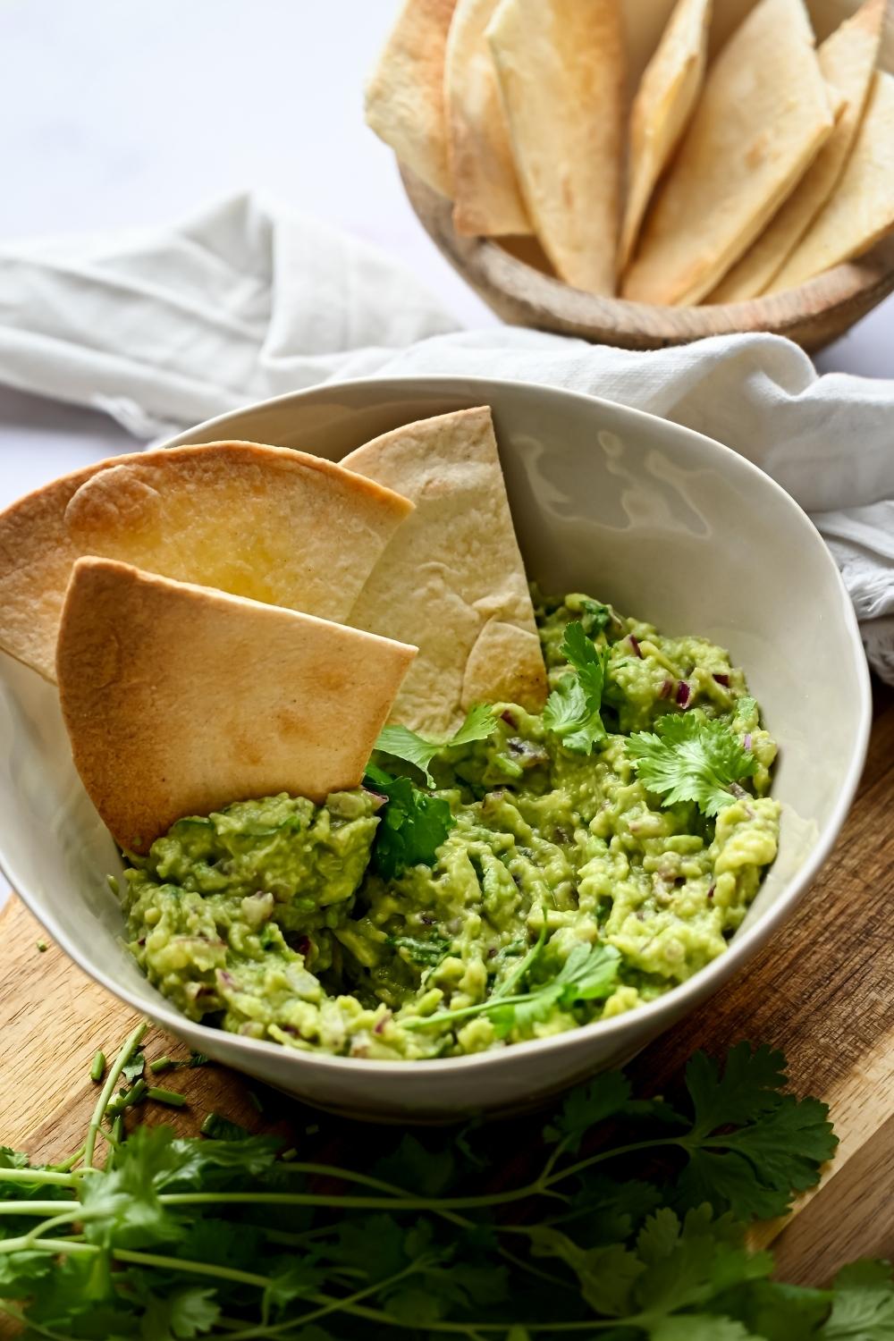 A serving bowl with homemade chipotle guac served on a wooden cutting board. A few tortilla chips are dipped in the bowl.