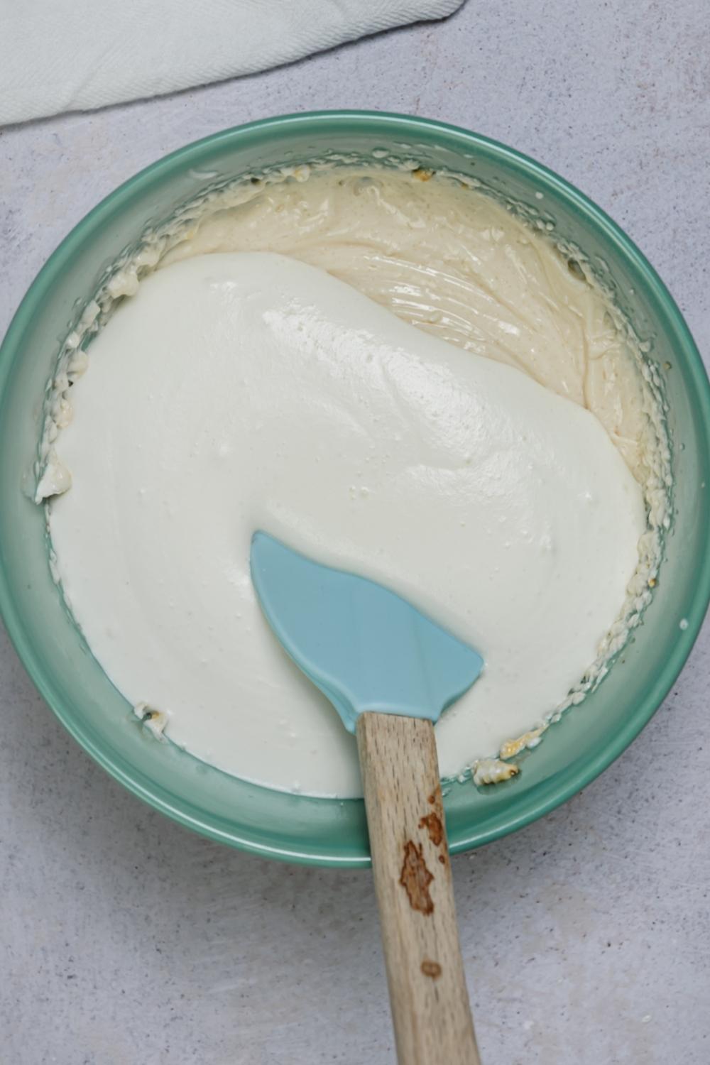 An overhead view of a mixing bowl with a creamy mixture of all the ingredients and cool whip on top. A rubber spatula sits in the bowl.