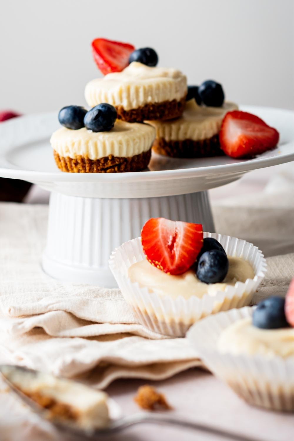 A cake stand with homemade mini cheesecake bites topped with berries. More mini cheesecake bites sit on the countertop in front of the cake stand. A spoon sits in front of those with a scoop of cheesecake in it.