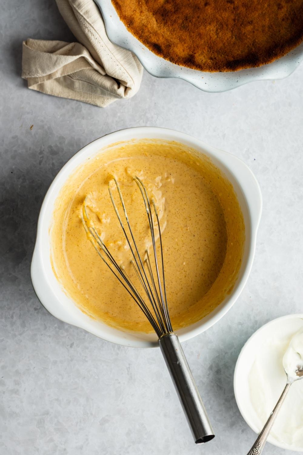 A whisk in a white bowl that is filled with mixed pumpkin pie ingredients.