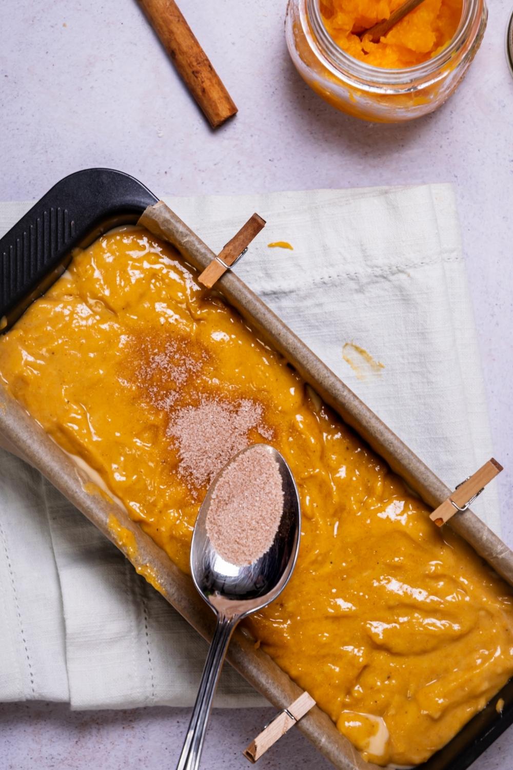 A spoon sprinkling cinnamon sugar on top of pumpkin bread batter in a loaf pan.