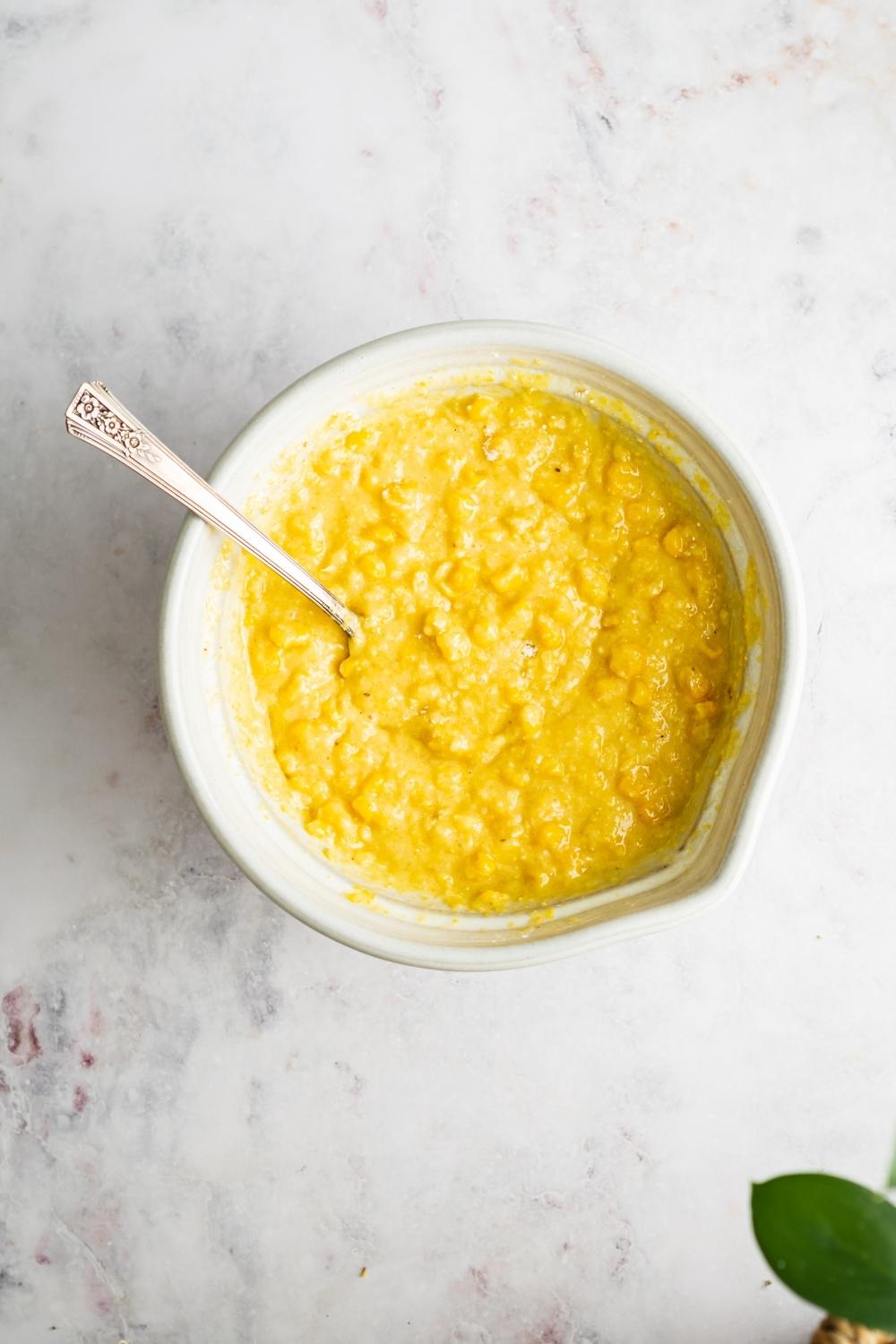A bowl that is filled with sweet corn cake batter with a spoon in it.