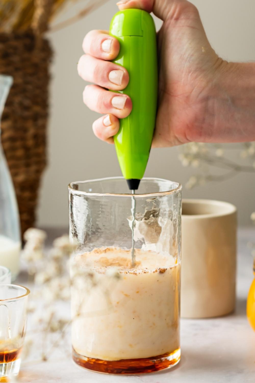 A hand holding an electric mixer, mixing pumpkin cream cold foam ingredients in a glass.
