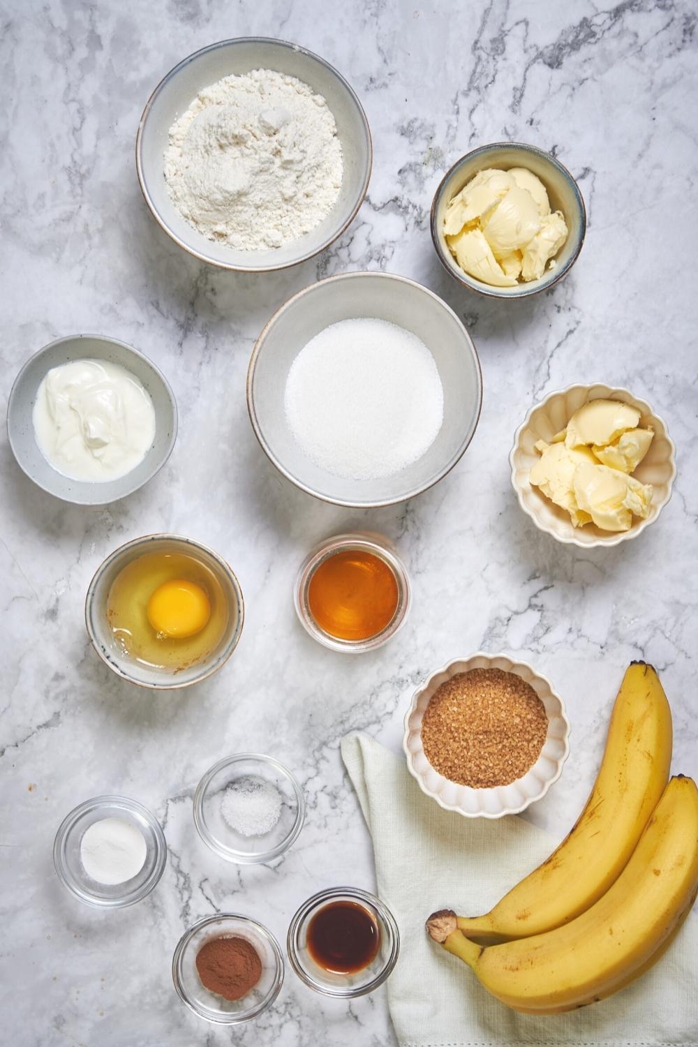 Bananas, a bowl of flour, a bowl of butter, a bowl of brown sugar, a bowl with an egg in it, a bowl of rum, and a bowl of vanilla on a grey counter.