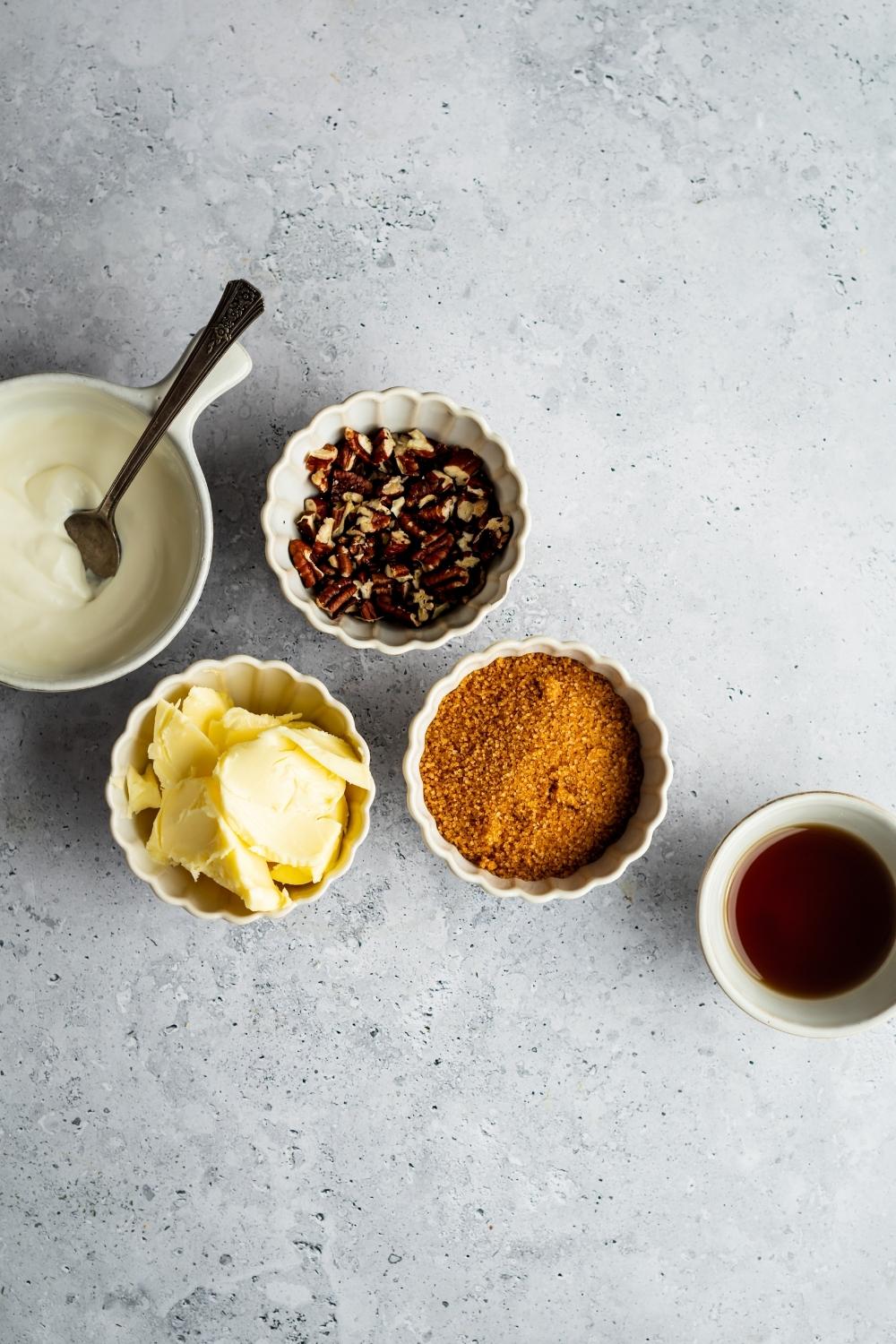 A bowl of heavy cream, a bowl of butter, bowl of chopped pecans, a bowl of brown sugar, and a bowl of maple syrup all on a gray counter.