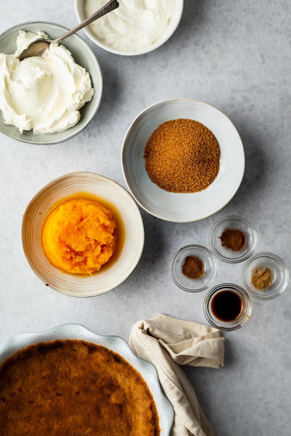 A bowl of pumpkin puree, a bowl of brown sugar, a bowl of vanilla extract, a bowl of cinnamon, a small bowl of nutmeg, a bowl of cream cheese, and part of a pie crust all on a grey counter.