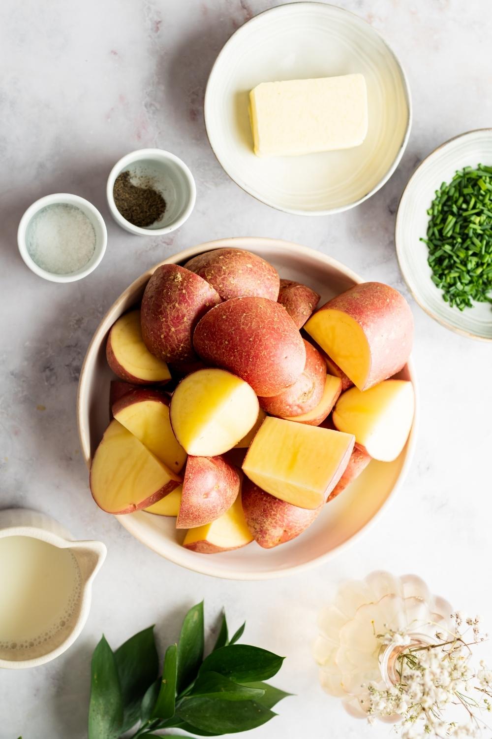 Sliced red skin potatoes in a bowl, a cube of butter on a plate, pepper in a small bowl, and salt in a small bowl all on a gray counter.