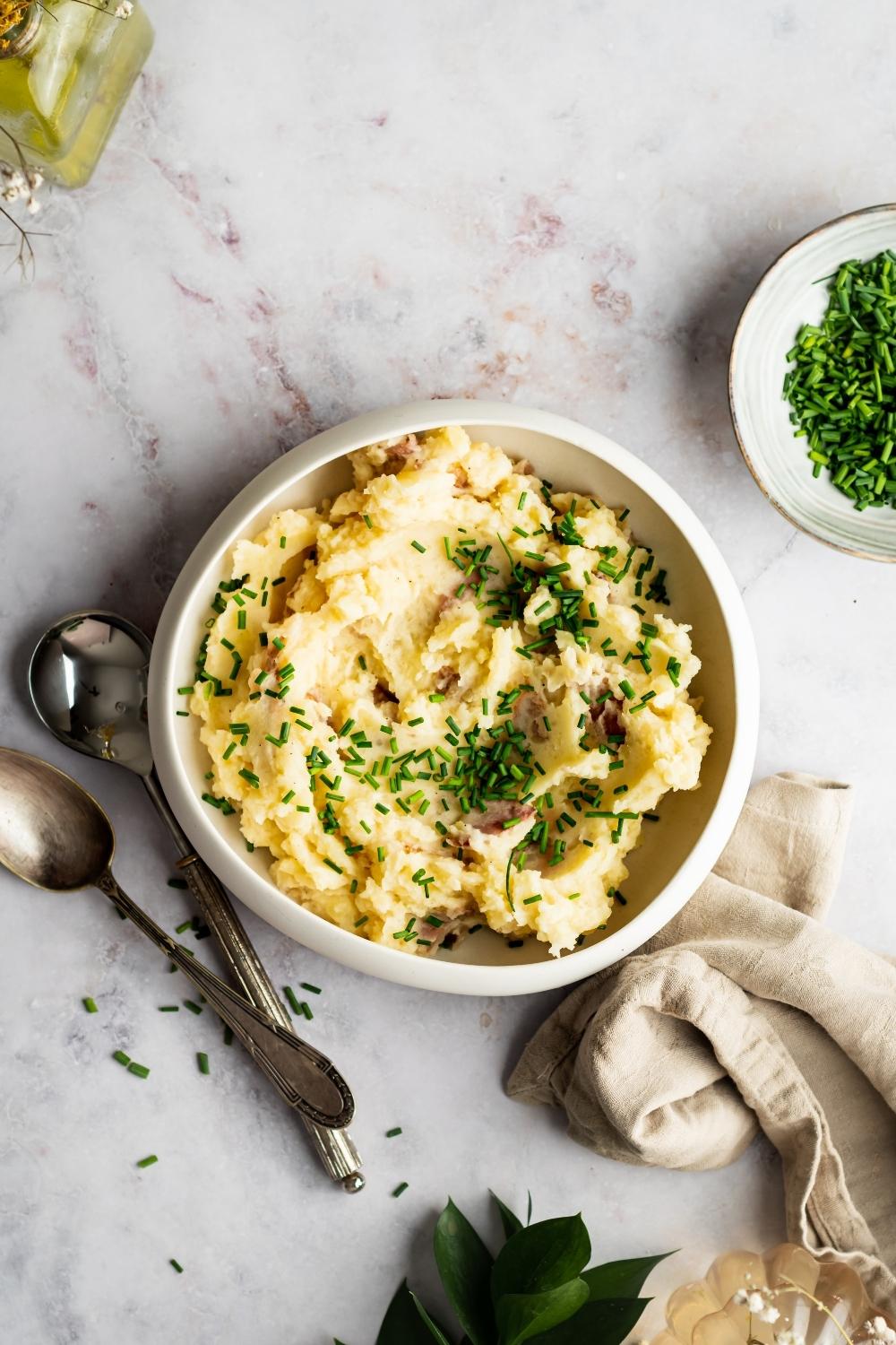 Red skinned mashed potatoes in a white bowl that is on a white counter with two spoons next to it and part of a plate of chopped green onion next to it.