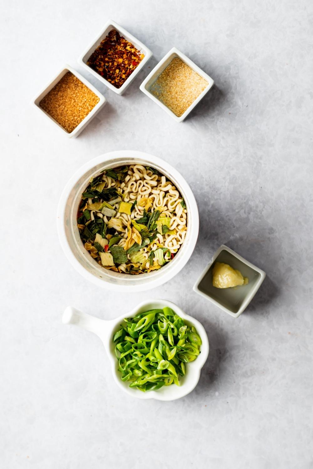 A bowl of chopped green onions, a bowl with butter in it, a bowl of ramen noodles, a square dish of red pepper flakes, a square dish of garlic powder on a grey counter.