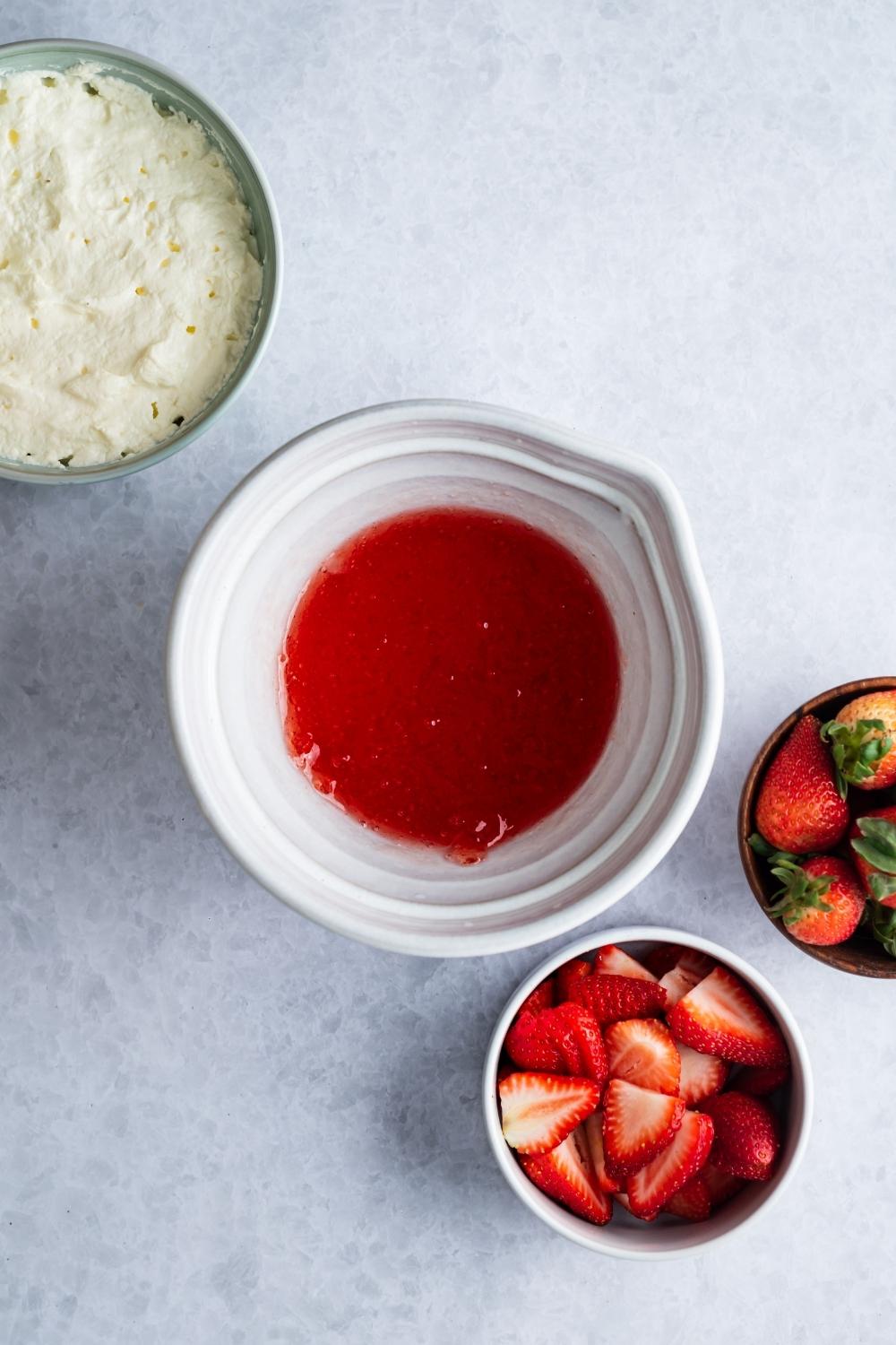Strawberry sauce in a white bowl. A bowl of sliced strawberries and cool whip are on the sides of it.