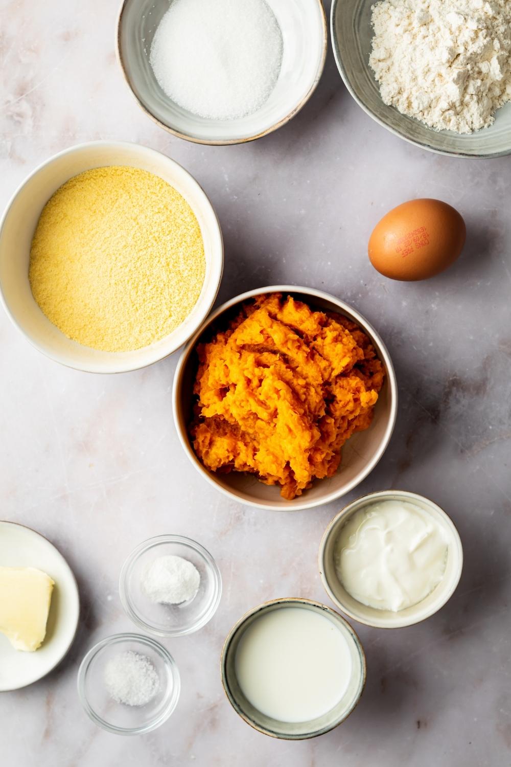 Mashed sweet potato in a bowl, egg, sour cream in a bowl, sugar in a bowl, cornmeal in a bowl, and part of a bowl of flour in it all on a gray counter.