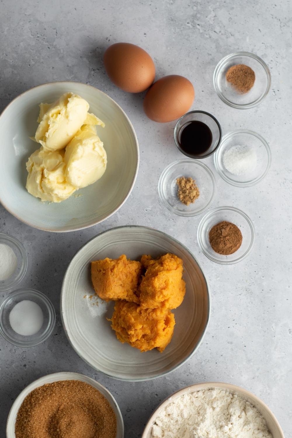 Sweet potato purée in a bowl, butter in a bowl, two eggs, a bowl of vanilla extract, bowl of ginger, bowl of cinnamon, but I'm not mad, both sugar, brown sugar, in part of a bowl of flour on a gray counter.