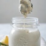 A small mason jar with homemade Alabama white sauce in it. A chicken wing has just been dipped into it and is suspended over top of the jar.