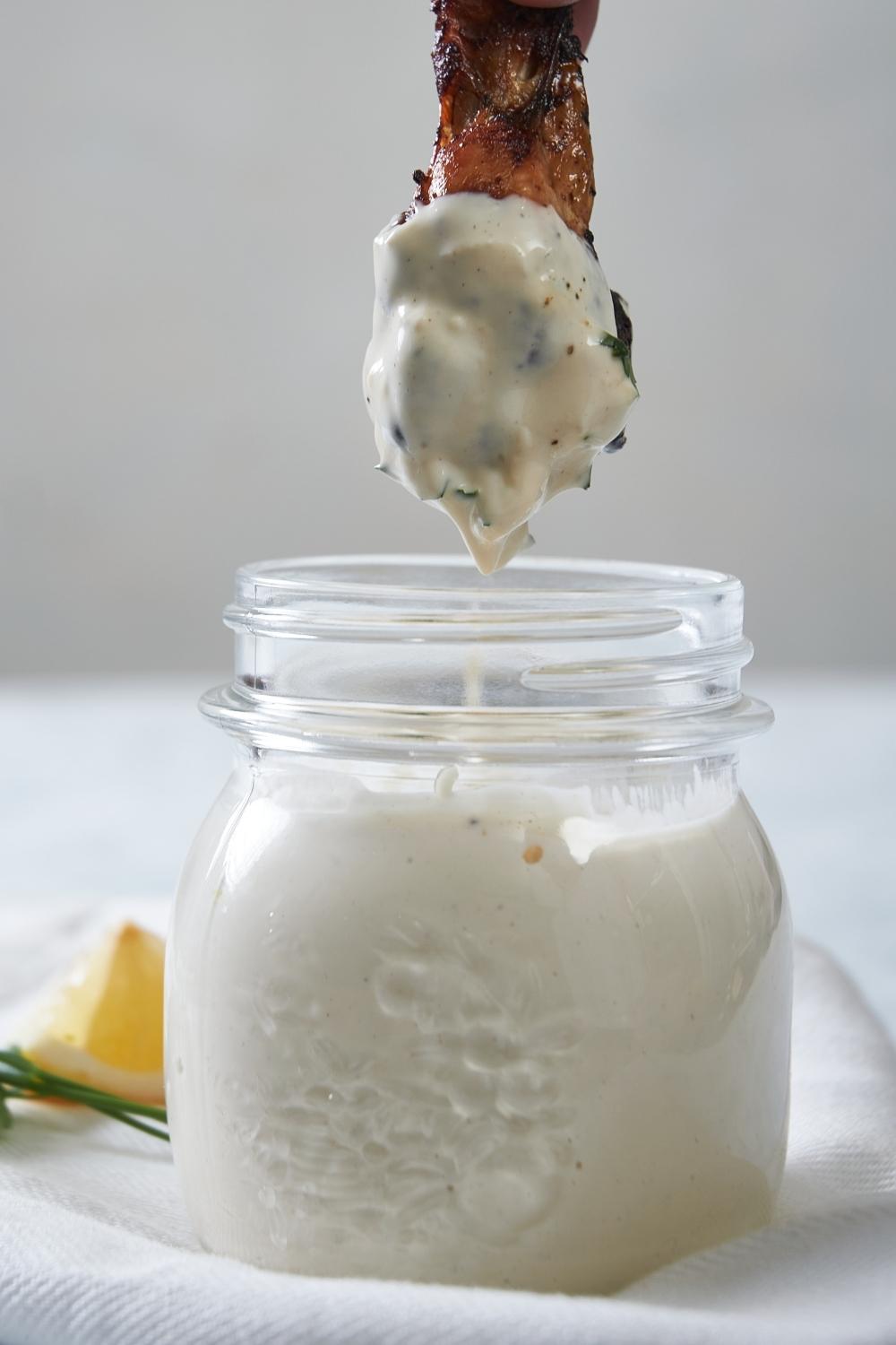A small mason jar with homemade Alabama white sauce in it. A chicken wing has just been dipped into it and is suspended over top of the jar.