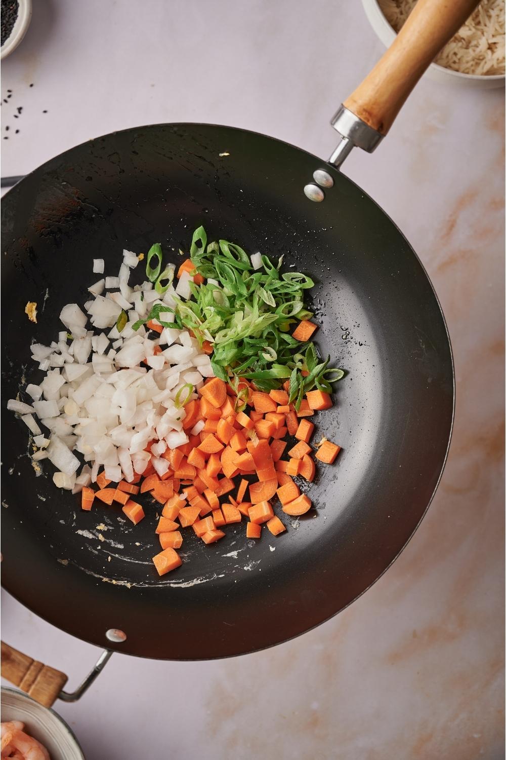 A wok on a grey counter with green onion, carrot, and white onion freshly added.