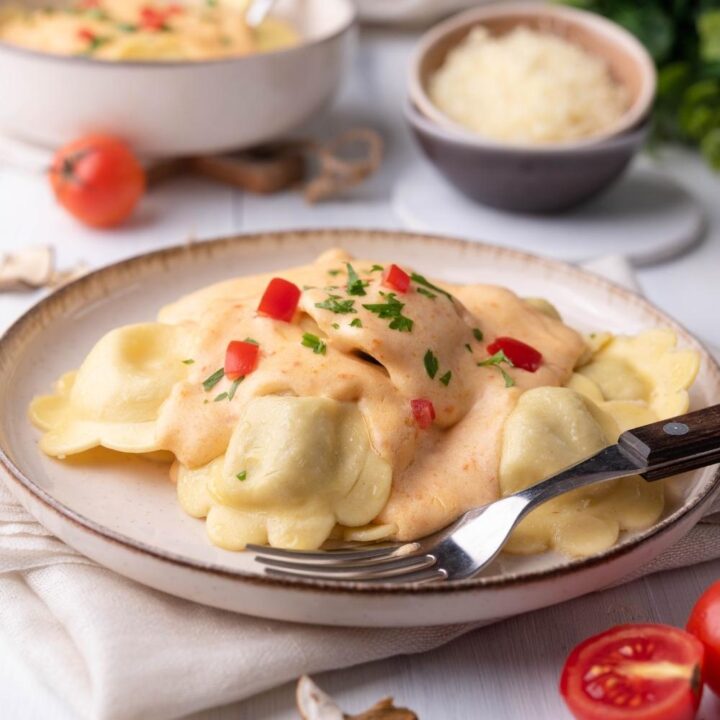Olive Garden cheese ravioli on a white plate with a fork on the plate. In the foreground there are cherry tomatoes, sliced mushrooms and a white kitchen towel. In the background there is a bunch of parsley, a bowl of shredded cheese, and a second bowl of pasta.