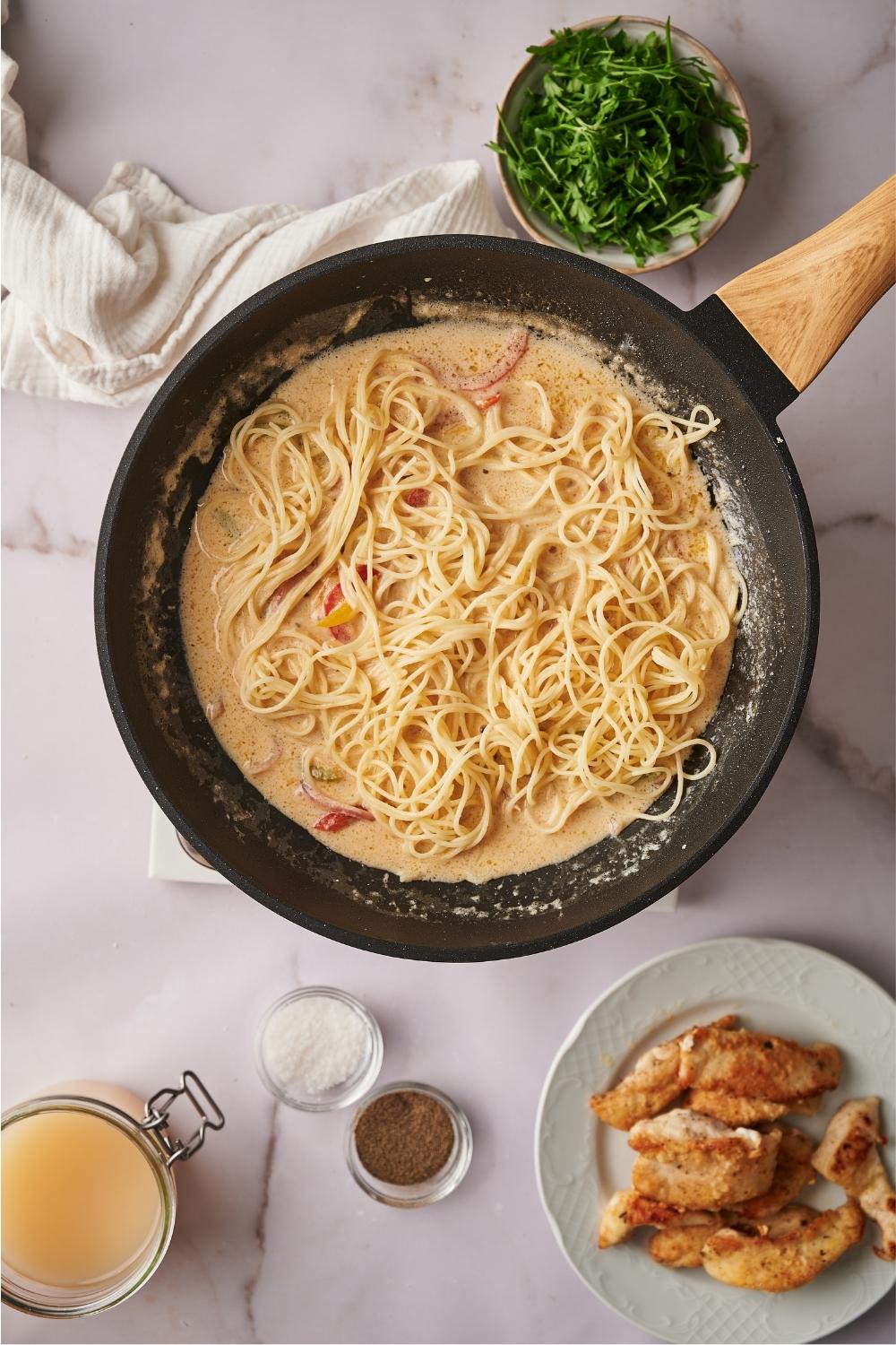 A skillet with sauteed veggies and scampi sauce with angel hair pasta freshly added to the skillet. Surrounding the skillet are plates and bowls of ingredients such as cooked chicken, salt, pepper, and parsley. There is a white napkin next to the skillet.