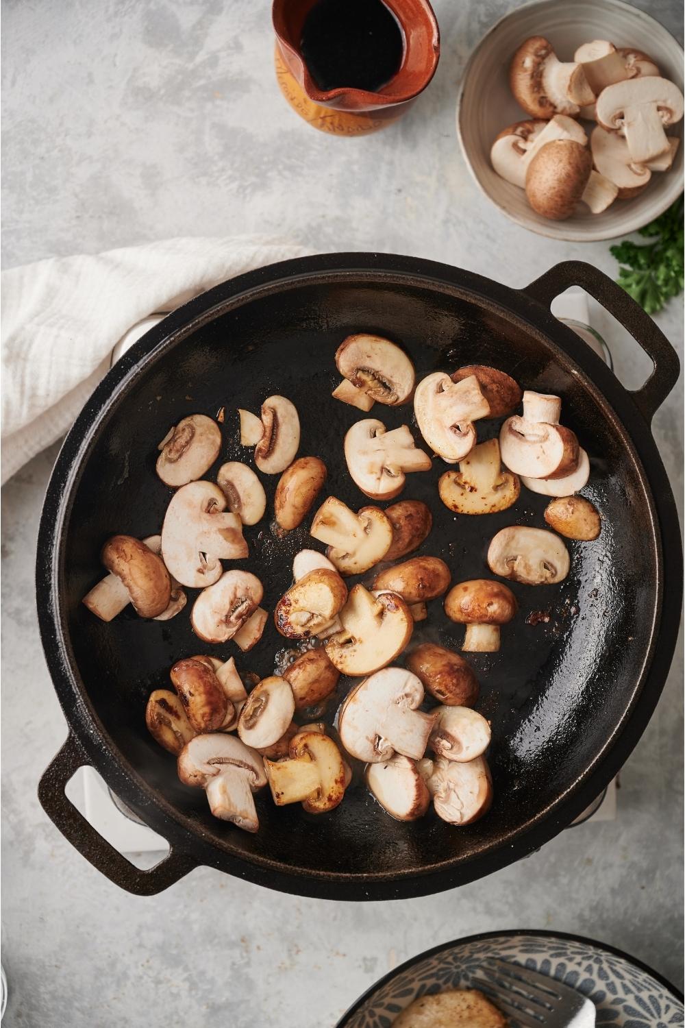 A cast iron pan with cooked and browned mushrooms in the pan, and a small bowl of raw mushrooms and marsala wine next to the pan.