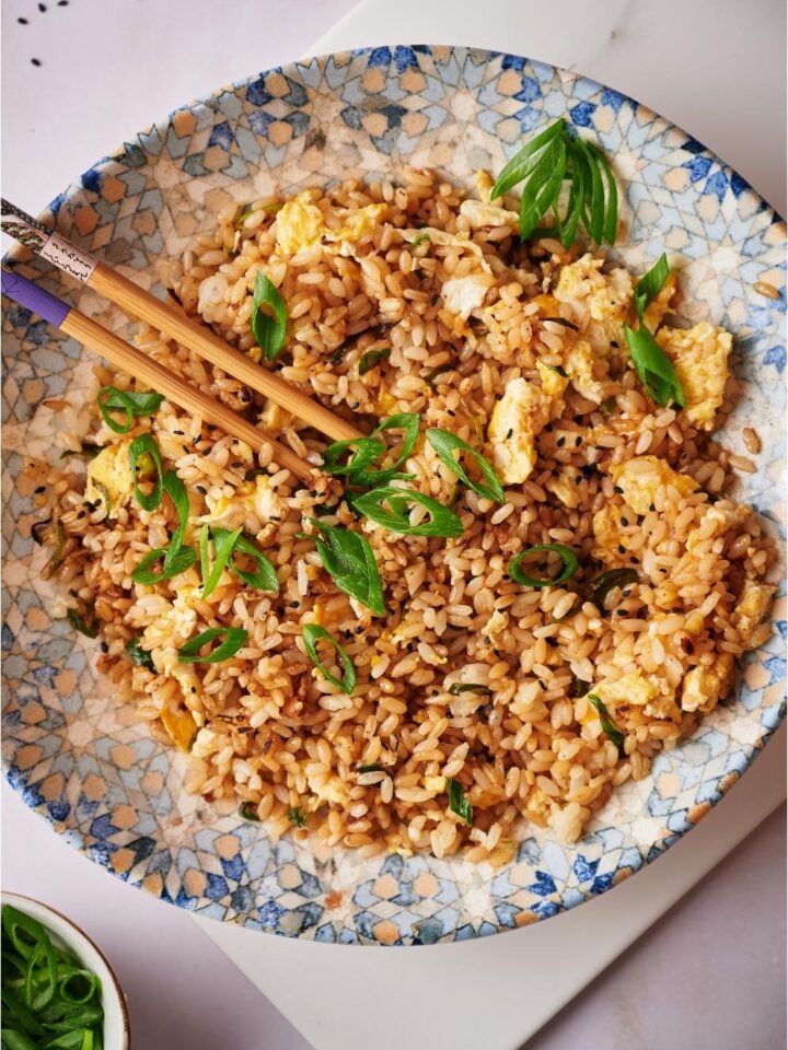 Hibachi fried rice in a decorated blue bowl with chopsticks, on a white board that is resting on a grey counter. In the corner there are sesame seeds and a bowl of chopped green onions.