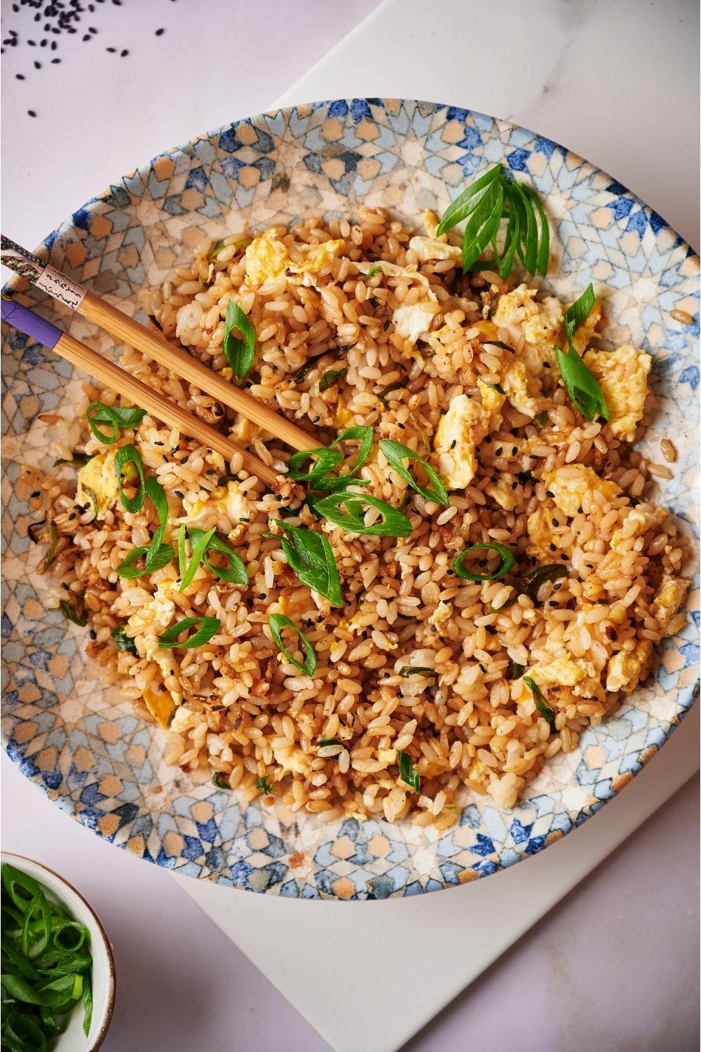 Hibachi fried rice in a decorated blue bowl with chopsticks, on a white board that is resting on a grey counter. In the corner there are sesame seeds and a bowl of chopped green onions.
