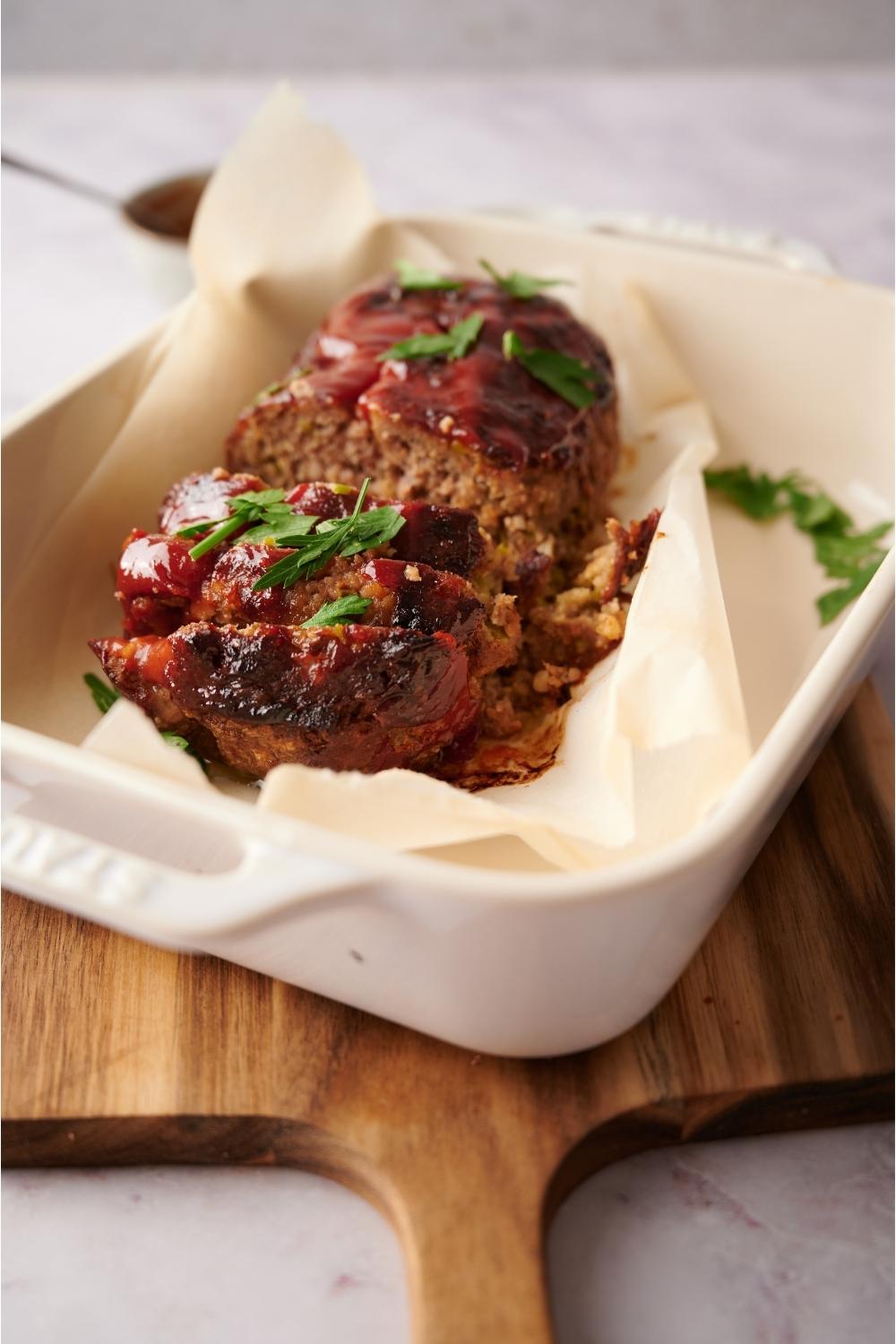 Southern meatloaf on parchment paper in a white baking dish. The dish is resting on a wood cutting board. Half of the meatloaf has been sliced to serve and the meatloaf is garnished with fresh parsley.