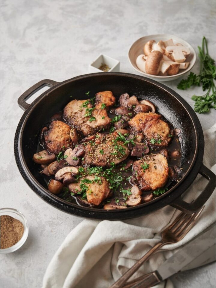 Cooked chicken marsala in a cast iron pan with a napkin, fork, knife, parsley sprigs, a bowl of raw mushrooms, and oregano all surrounding the pan on a grey counter.