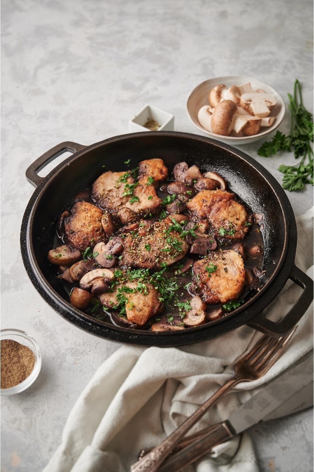 Cooked chicken marsala in a cast iron pan with a napkin, fork, knife, parsley sprigs, a bowl of raw mushrooms, and oregano all surrounding the pan on a grey counter.