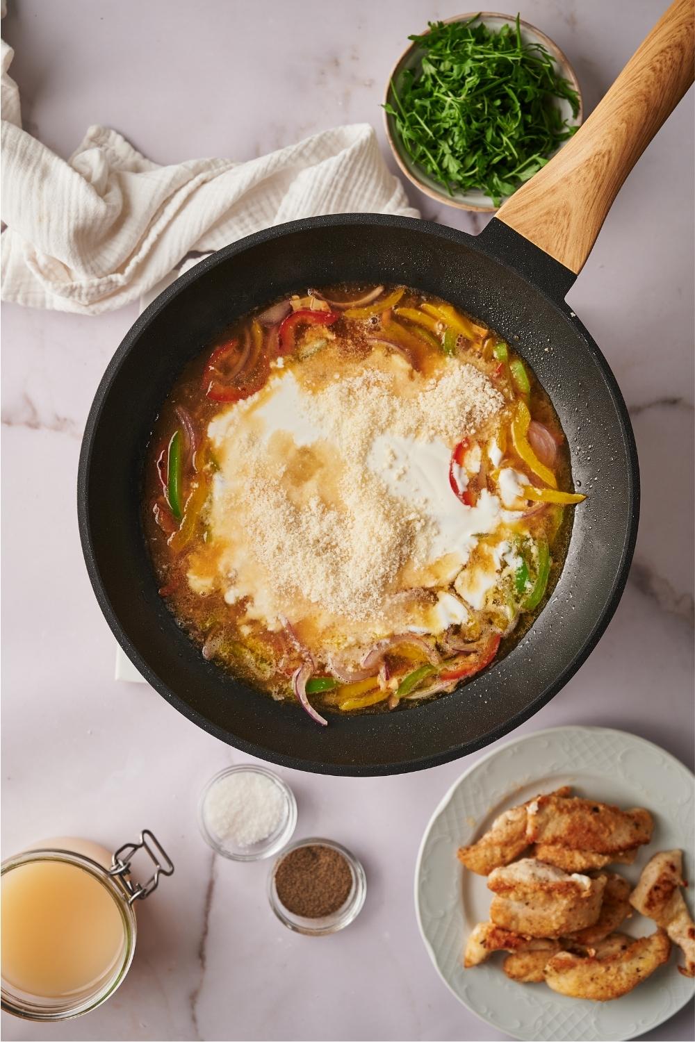 A skillet containing cooked bell pepper and red onion with scampi sauce ingredients and parmesan cheese freshly added to the skillet. Surrounding the skillet are plates and bowls containing ingredients such as cooked chicken, salt, pepper, and parsley.
