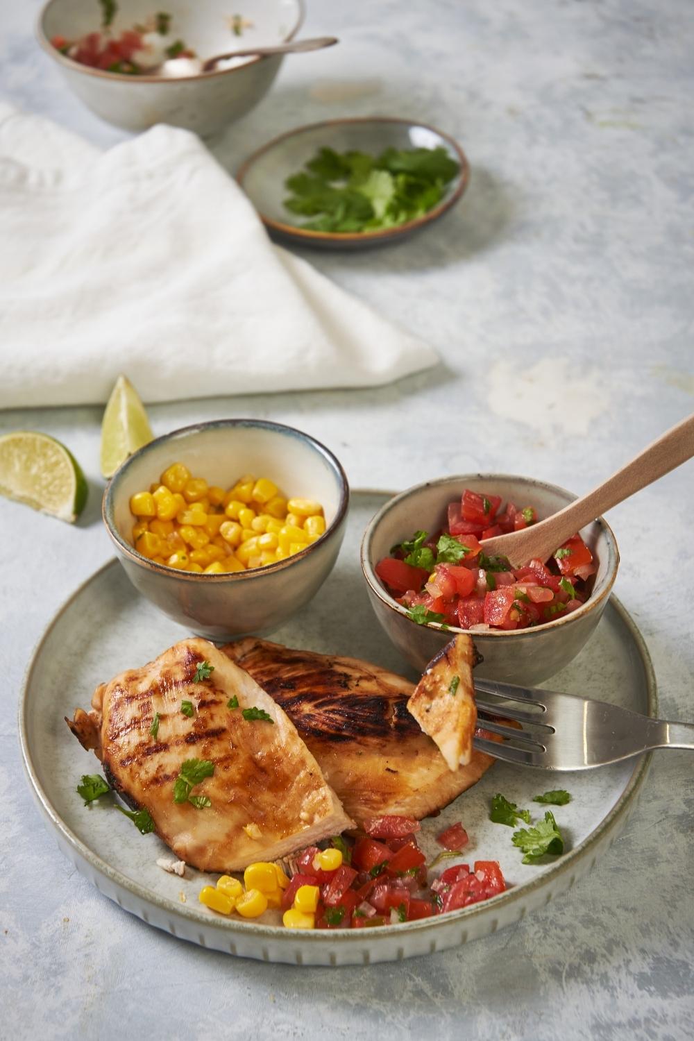 Chili's margarita chicken on a grey plate on a grey counter, with a fork grabbing a piece of chicken and containers of pico and corn are on the plate. There is a spoon in the pico and an assortment of ingredients behind the plate.