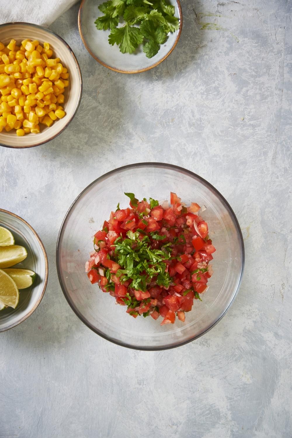 Clear bowl of pico de gallo ingredients on a grey counter next to bowls of corn, cilantro, and lime wedges.