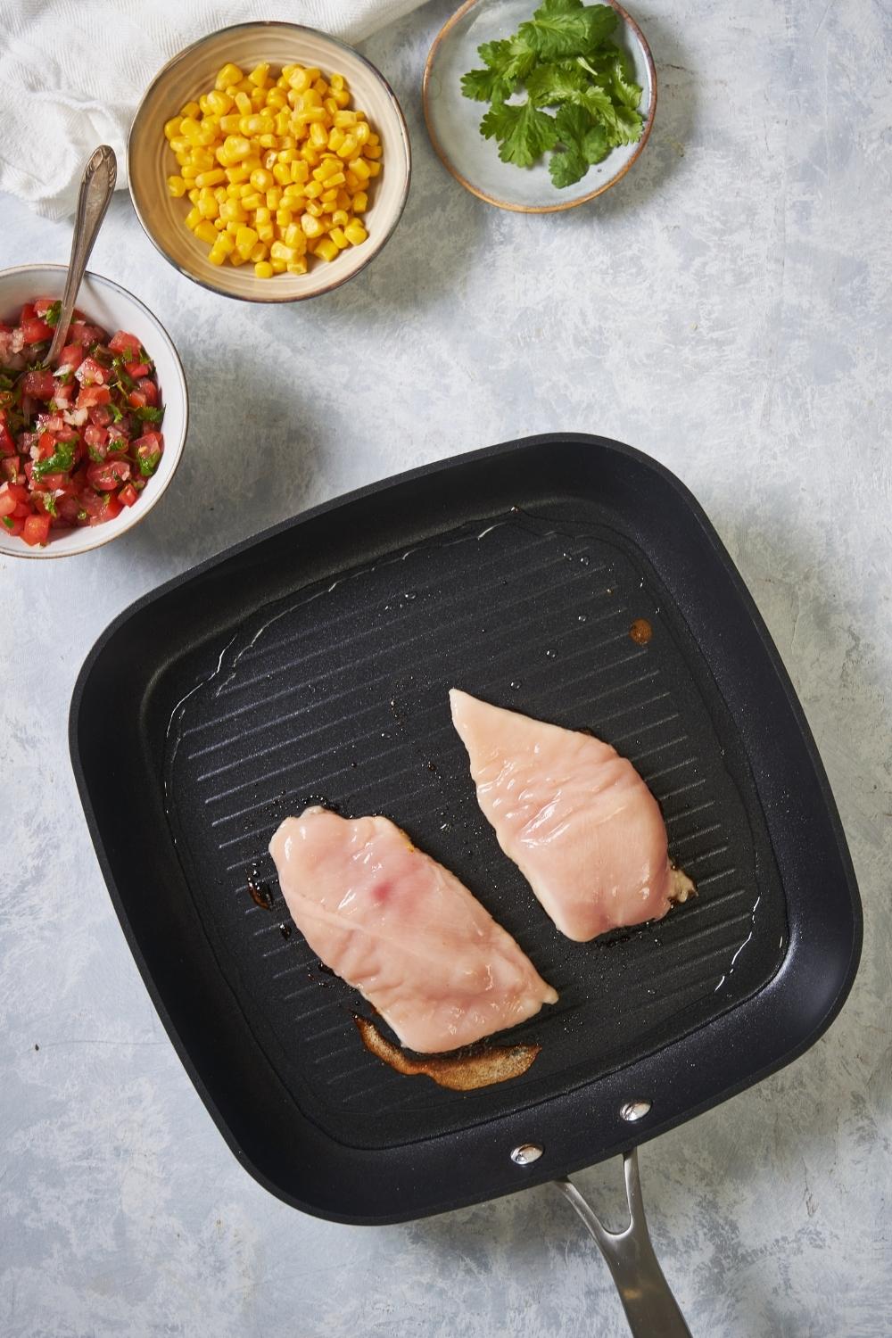 A grill pan on a grey counter with raw chicken breasts, surrounded by bowls of corn, pico, and cilantro.