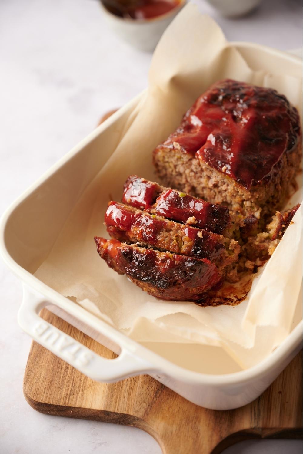 Southern meatloaf on parchment paper in a white baking dish. The dish is resting on a wood cutting board. The meatloaf has been slice to serve and layers are stacked on top of one another.